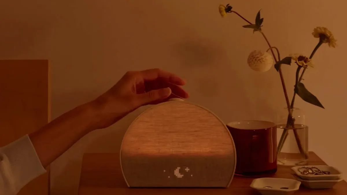 Mão tocando o relógio Hatch, que simula o amanhecer, sobre uma mesa de cabeceira com luz âmbar e um símbolo de lua no visor