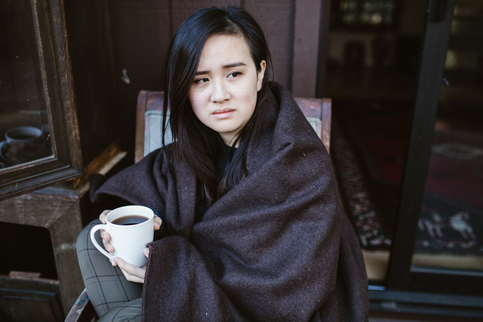 Mulher envolta em um cobertor segurando uma xícara de café, olhar pensativo e introspectivo